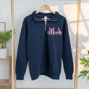 May include: Navy blue sweatshirt with a quarter-zip collar. The sweatshirt features the name "Alexis" in a cursive font, with "MA" above it, in pink. The long-sleeved top is hanging on a wooden clothes rack.