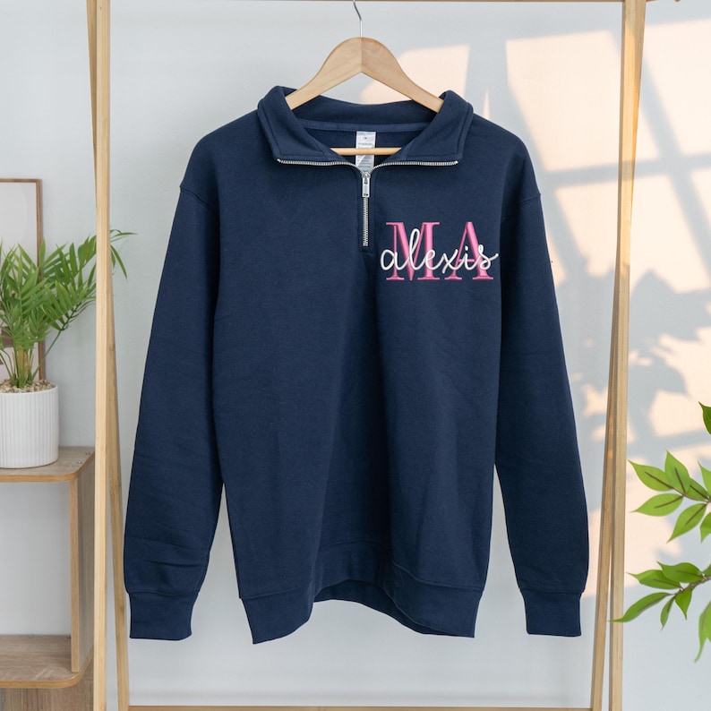 May include: Navy blue sweatshirt with a quarter-zip collar. The sweatshirt features the name "Alexis" in a cursive font, with "MA" above it, in pink. The long-sleeved top is hanging on a wooden clothes rack.