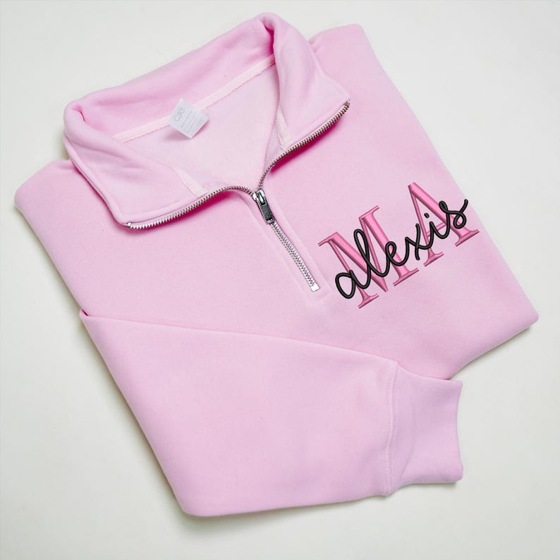 May include: A pale pink quarter-zip sweatshirt featuring the name "Alexis" embroidered in pink and black. The sweatshirt has a folded collar and a silver zip. The fabric appears soft and comfortable.