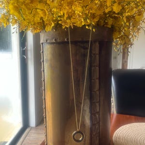 May include: A gold necklace with a round pendant hangs in a rustic metal vase filled with yellow flowers.