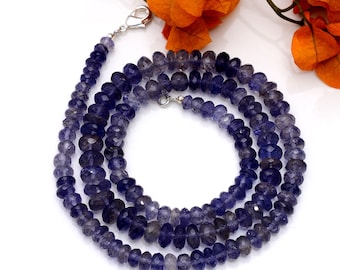 May include: A strand of dark blue gemstone beads, likely amethyst, strung on a silver chain. The beads are faceted and have a slightly irregular shape. The necklace is displayed on a white surface with orange flowers in the background.