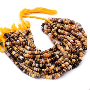 Könnte beinhalten: Eine Kette aus kleinen, runden Perlen in Braun-, Beige- und Cremetönen. Die Perlen sind auf einer gelben Schnur mit gelben Quasten aufgefädelt. Die Perlen sind gleichmäßig groß und geformt und erzeugen eine natürliche, erdige Ästhetik.