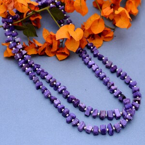 May include: A double strand necklace made of purple and silver square-shaped beads. The necklace is lying on a blue surface with orange flowers behind it.