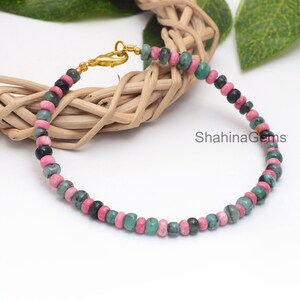 May include: A beaded bracelet featuring a mix of pink, green, and black gemstones. The bracelet is strung on a gold-colored chain and clasp. The gemstones are round and evenly spaced.