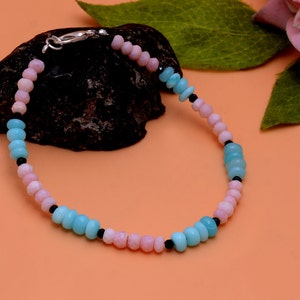 Könnte beinhalten: Ein zartes Armband mit abwechselnd hellrosa und hellblauen Perlen, durchsetzt mit kleinen schwarzen Perlen. Das Armband wird mit einem silbernen Verschluss befestigt und wird auf einer dunklen, strukturierten Oberfläche präsentiert.