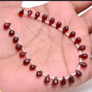 Puede incluir: Hilo de cuentas de granate rojo oscuro, cada una tallada en forma de lágrima facetada. Las cuentas están ensartadas en un fino alambre plateado. Las cuentas tienen una superficie pulida y varían ligeramente en tamaño. Aproximadamente 0,5 cm de largo.