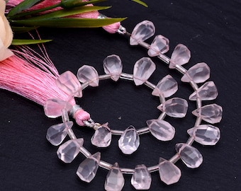 May include: Two strands of faceted, light pink rose quartz teardrop beads. The beads are strung on a clear wire. A pink tassel and faux greenery are in the background.