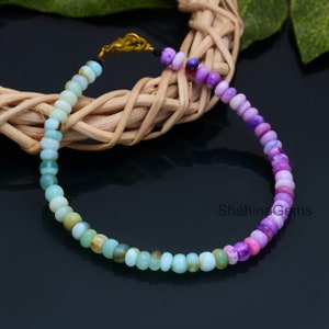 May include: A beaded bracelet featuring a gradient of colors, transitioning from light blue and green to purple and pink. The bracelet is made of small, round beads and has a gold-colored clasp. The beads are strung together to form a flexible band.