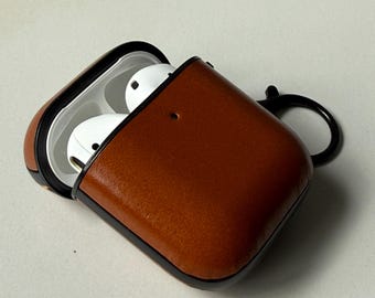 May include: A brown leather protective case for wireless earbuds, featuring a smooth finish, a black trim, and a black circular carabiner clip attached to the side. The case is open, revealing white wireless earbuds nestled inside the white interior.
