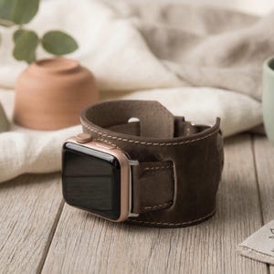 Könnte beinhalten: Ein braunes Lederarmband für die Apple Watch mit einem goldfarbenen Zifferblatt. Das Armband ist mit weißem Faden genäht und hat einen Schnallenverschluss. Die Uhr wird auf einer Holzoberfläche präsentiert.