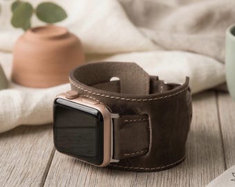May include: A brown leather Apple Watch band with a gold-colored watch face. The band is stitched with white thread and has a buckle closure. The watch is displayed on a wooden surface.