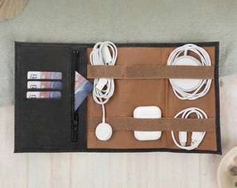 May include: Black and brown leather tech organizer with multiple compartments. Features a zippered pocket, card slots, and elastic straps to hold white charging cables, a charging pad, and wireless earbuds. Ideal for travel.