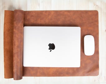 May include: A brown leather desk mat with a laptop and a white mouse. The laptop is silver with a black apple logo. The desk mat is rectangular, with a rolled-up edge on the left side.