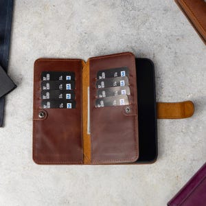 May include: A brown leather wallet case for a smartphone, featuring an open design with multiple card slots on the left and right interior panels. The case includes a snap button closure and holds several black credit cards with visible Visa logos.