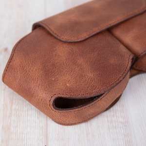 K&ouml;nnte beinhalten: Eine braune Ledertasche mit einem Klappverschluss und einem gen&auml;hten Design. Die Tasche hat eine abgerundete Form und eine glatte Textur.