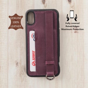 May include: A burgundy leather phone case with a black frame. The case features a card slot with a white and red card, a wrist strap, and a metal ring. Text on the case reads "Fully covered, Raised Edges, Maximum Protection" and "Genuine 100% Leather."