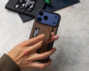 Puede incluir: Una funda de cuero marrón oscuro para teléfono con ranura para tarjetas integrada y una correa vertical para agarre. La funda tiene un recorte para un sistema de cámara de triple lente y sostiene una tarjeta negra con el texto CREDIT CARD.