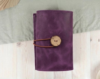 May include: A deep purple leather wallet with a wooden button closure and a brown leather strap. The wallet is closed and has a textured surface. The background is a light wood surface with a green cloth.
