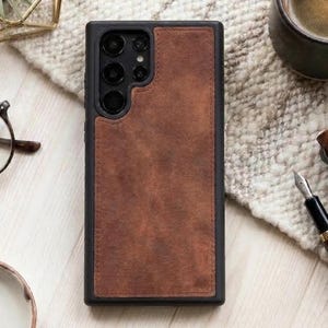 May include: A black smartphone case with a brown leather panel. The case has a rectangular shape with a camera cutout and stitched edges. The leather panel is a warm brown color, and the case is on a textured surface.