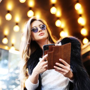 K&ouml;nnte beinhalten: Eine Frau mit Sonnenbrille und Pelzmantel h&auml;lt eine braune Leder-Handyh&uuml;lle mit einem schwarzen Smartphone darin.