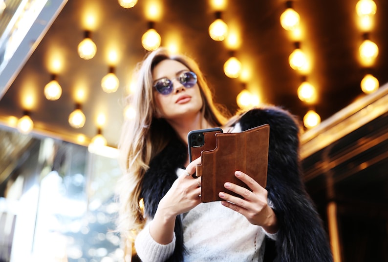 K&ouml;nnte beinhalten: Eine Frau mit Sonnenbrille und Pelzmantel h&auml;lt eine braune Leder-Handyh&uuml;lle mit einem schwarzen Smartphone darin.