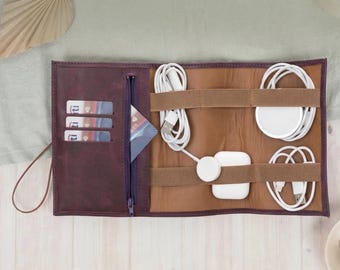 May include: A burgundy leather tech organizer, open to display white charging cables, a wireless charger, and a smartwatch charger. The interior is brown with elastic straps. A zippered pocket and card slots are visible.