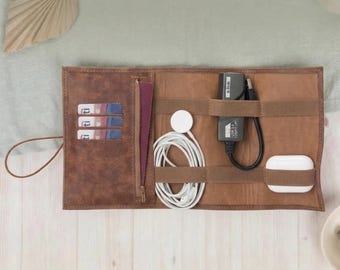 May include: A brown leather travel organizer, open to display its contents. It features slots for cards, a zippered pocket, and elastic straps holding a white charging cable, a black USB adapter, a white smartwatch charger, and a white wireless earbud case.