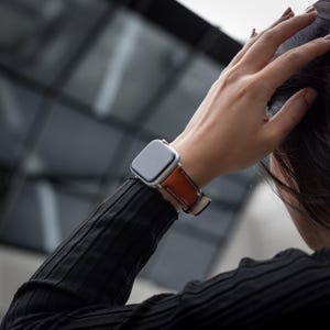 May include: A silver Apple Watch with a brown leather band. The watch is on a person's wrist.