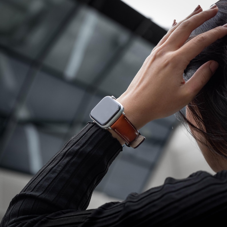 May include: A silver Apple Watch with a brown leather band. The watch is on a person's wrist.