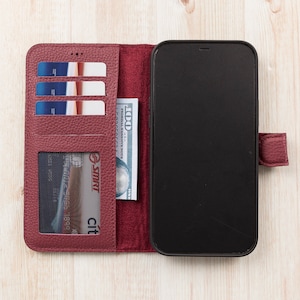 K&ouml;nnte beinhalten: Ein burgunderfarbenes Leder-Handy-Etui mit integriertem Portemonnaie. Das Etui hat F&auml;cher f&uuml;r Kreditkarten und Bargeld. Das Handy-Etui ist ge&ouml;ffnet und ein schwarzes Smartphone ist sichtbar.