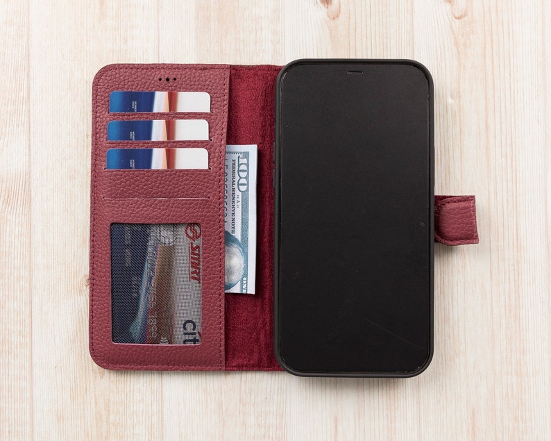 K&ouml;nnte beinhalten: Ein burgunderfarbenes Leder-Handy-Etui mit integriertem Portemonnaie. Das Etui hat F&auml;cher f&uuml;r Kreditkarten und Bargeld. Das Handy-Etui ist ge&ouml;ffnet und ein schwarzes Smartphone ist sichtbar.
