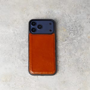 May include: A smartphone case featuring a dark gray frame and a smooth, tan-colored leather back panel. The upper section of the case is dark gray and includes cutouts for a triple-lens camera system, a flash, and a sensor array.