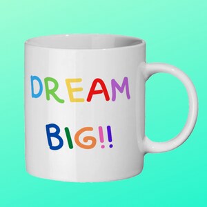 May include: White ceramic mug featuring the phrase "DREAM BIG!!" in vibrant, multi-coloured letters. The mug has a curved handle and a glossy finish, suitable for hot beverages like tea or coffee.