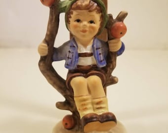 May include: A porcelain figurine of a boy sitting on a tree branch with apples. He is wearing a blue and white shirt, brown pants, and a green hat. The figurine is about 4 inches tall.