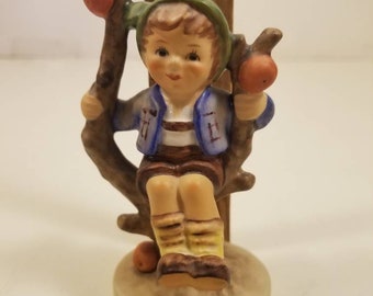 May include: A porcelain figurine of a boy sitting on a tree branch with apples. He is wearing a blue jacket, brown shorts, and a green hat. The figurine is about 4 inches tall.