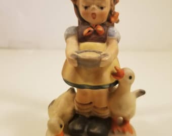 May include: A porcelain figurine of a girl with closed eyes holding a bowl, surrounded by two white geese. The girl is wearing a brown dress with a yellow apron and a blue and white collar.