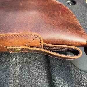 May include: A brown leather gun case with a strap and a brass button. The case is lying on a black textured surface.
