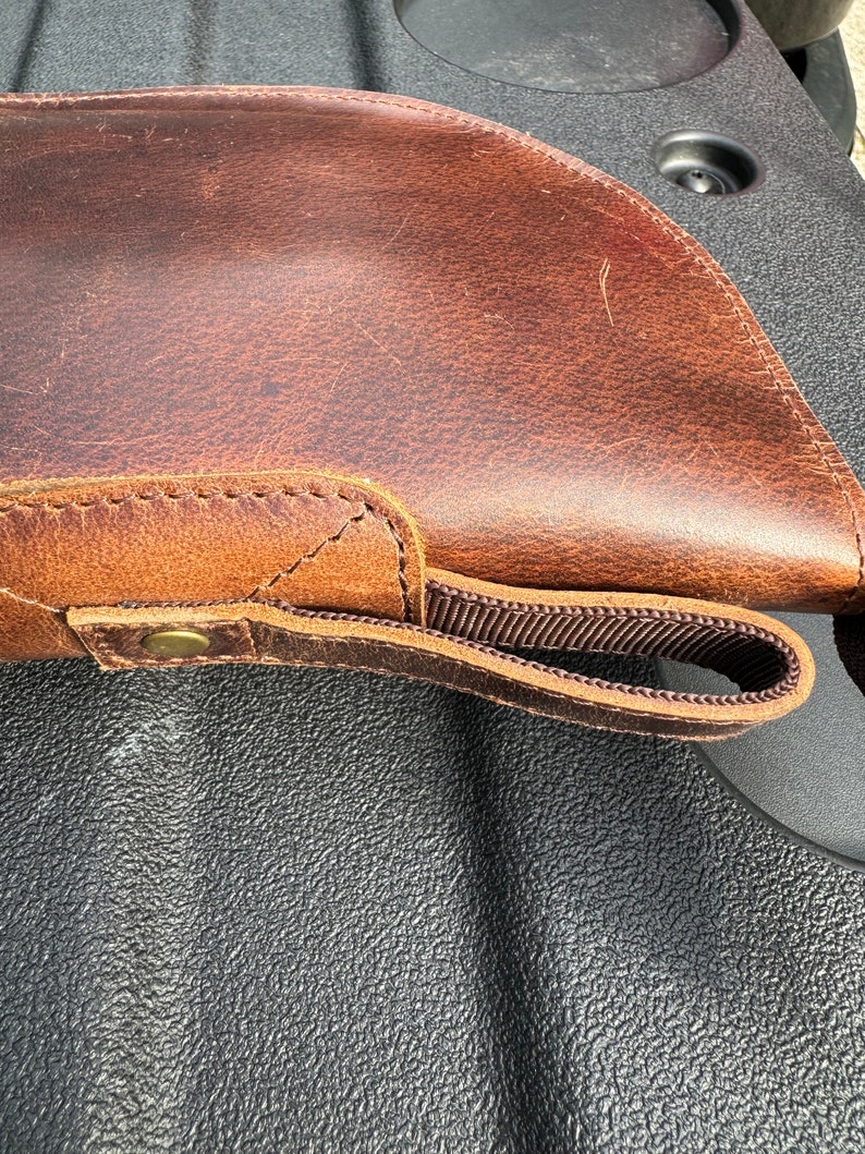 May include: A brown leather gun case with a strap and a brass button. The case is lying on a black textured surface.