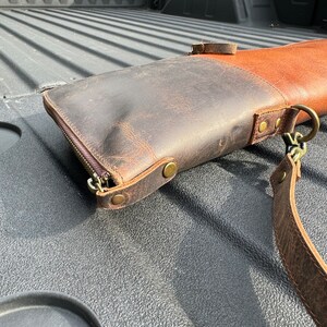 May include: A brown leather gun case with a zipper closure and a strap. The case is lying in the bed of a pickup truck.