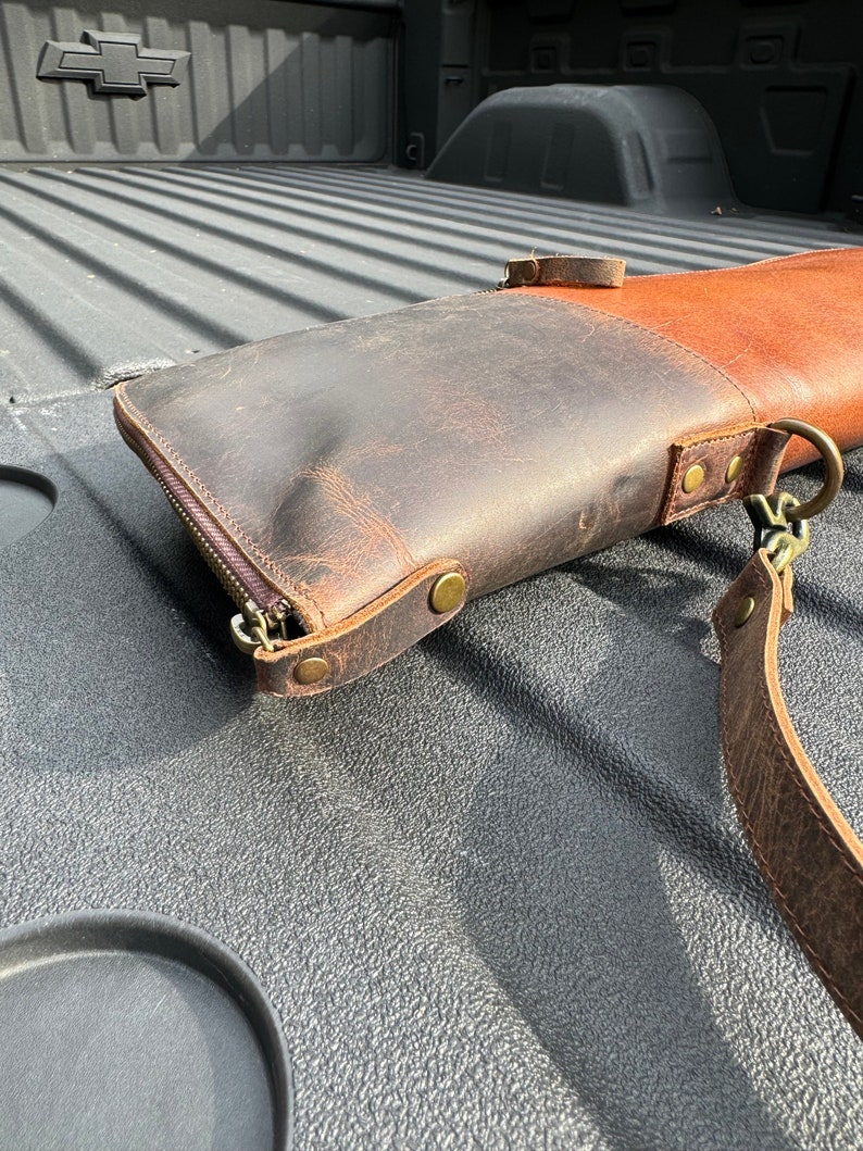 May include: A brown leather gun case with a zipper closure and a strap. The case is lying in the bed of a pickup truck.