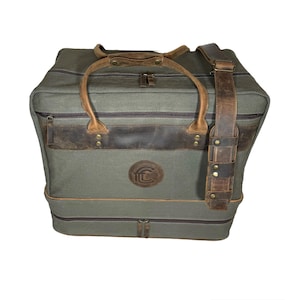 May include: A large, olive green travel bag with brown leather accents. The bag features a top handle, a detachable shoulder strap, and a circular logo. The bag has a zippered top and a zippered bottom compartment.