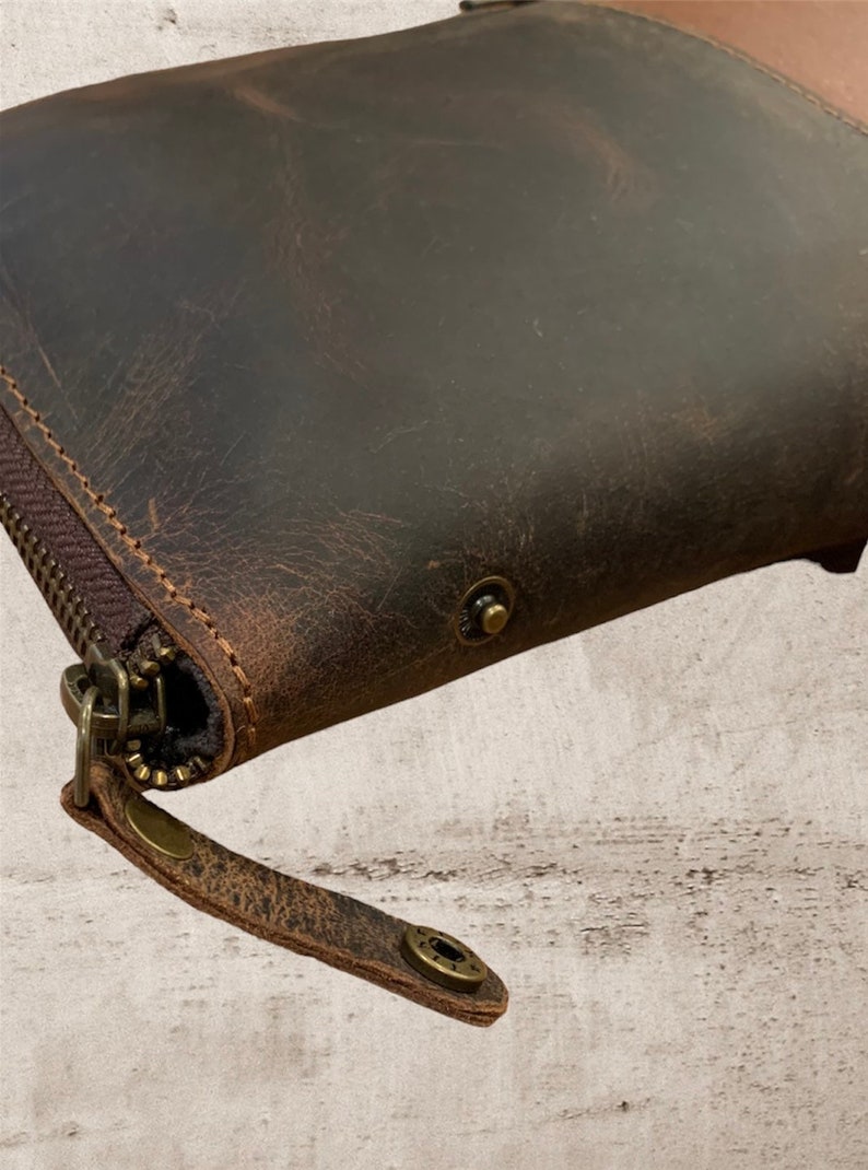 May include: A brown leather wallet with a zipper closure and a brown leather strap. The wallet has a distressed look and features brass accents.