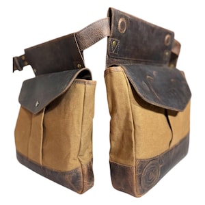 May include: A pair of brown and tan leather and canvas bags. The bags have a flap closure and are attached to a leather belt. The bags have a distressed leather trim and a logo on the bottom.
