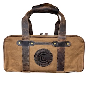 May include: A brown canvas and leather duffel bag with a circular logo featuring the letters "FTC" in a black circle. The bag has two brown leather handles and a zipper closure.