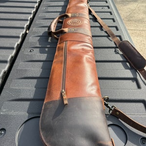 May include: A brown leather gun case with a black leather bottom and a zipper closure. The case has a shoulder strap and a handle.