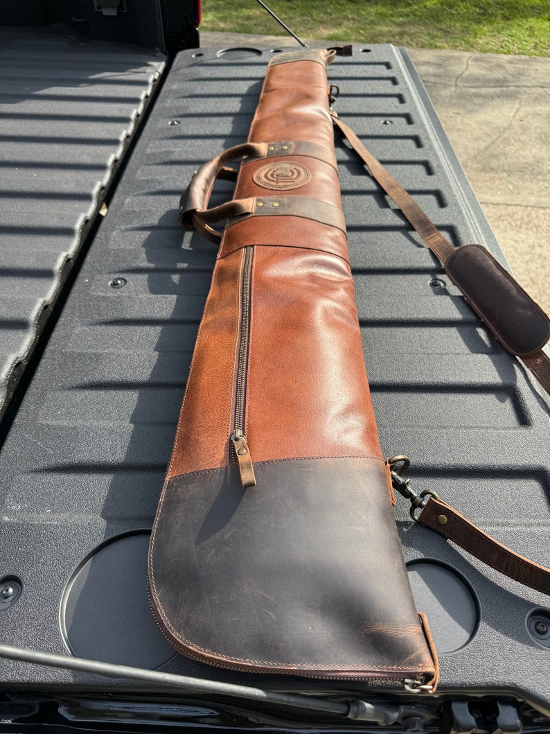 May include: A brown leather gun case with a black leather bottom and a zipper closure. The case has a shoulder strap and a handle.