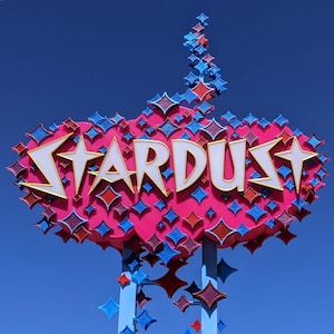 May include: A large, pink, cloud-shaped sign featuring the word STARDUST in white, stylized, gold-bordered letters. The sign is decorated with numerous four-pointed star shapes in shades of blue and red, mounted on two light blue vertical poles.
