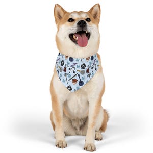 May include: A Shiba Inu dog wearing a light blue bandana with a repeating pattern of various symbols, including a paw print, lightning bolt, and a hammer. The dog is sitting and looking up with its tongue out.