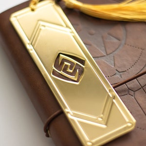 May include: A brown leather journal with a gold bookmark featuring a geometric design. The bookmark has a tassel made of yellow yarn.