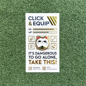 May include: Gold-coloured enamel pin featuring a white cat face with red cheeks and closed eyes. The pin is on a white card with the text "CLICK & EQUIP" and "IT'S DANGEROUS TO GO ALONE, TAKE THIS!" in gold lettering.